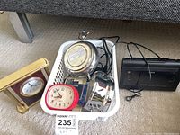 Lot of clocks and basket on carpet, showing multiple clocks including a brass clock, red alarm clock, round silver twin bell clock, and black digital clock radio.