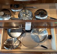 Top-down view in drawer showing several stainless steel pots and pans with lids, including the perforated steamer insert and fry pan.