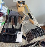 Photo of various knives laid out on a surface alongside a knife block and a knife sharpener tool.