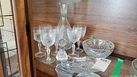 Photo showing decanter, six stemmed glasses, large bowl, and serving plate on a glass shelf.