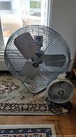 Two Patton fans positioned beside each other on a carpet. Larger floor fan with a metal cage and blades, smaller desk fan in front.