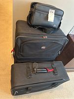 Photo showing three black luggage pieces stacked in a corner: the largest suitcase, a medium-sized suitcase with buckled straps, and a small travel bag on top.
