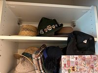 Photo showing a shelf with various men's hats including straw hats, a plaid newsboy cap, and a bucket hat with other soft fabric hats in the background.