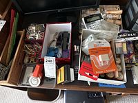 Top-down view of cardboard box with assorted small office supplies, calculator, batteries, and rolls of clear adhesive tape.