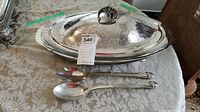 Oval silverplate serving dish with decorated lid, glass insert inside, and two serving utensils laid in front on a patterned tablecloth.