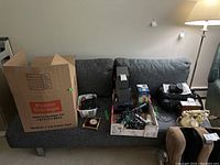 Wide view of grey sofa bed with removable back cushions. Sofa has grey woven fabric with tight weave texture. Multiple boxes and various household items placed on top.