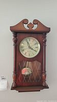 Full view of the wooden Daniel Dakota wall clock showing the ornate top and turned side columns, clock face, and glass pendulum door.