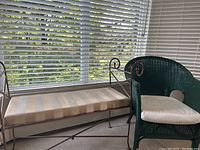 Green wicker club chair with white cushion and metal bench with striped padded seat, placed indoors near window with blinds.
