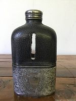 Front view of antique flask showing embossed silver plate with shield emblem and black leather covering glass body.