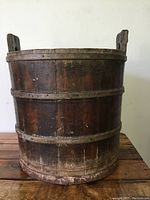 Front view of an antique wooden bucket made of vertical wooden slats and three horizontal bands, showing rustic wear.