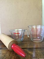Wooden rolling pin with red painted handle and two Pyrex measuring cups showing the items and condition.