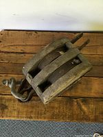Top angle view showing front and side of the antique wooden pulley resting on a wooden surface, highlighting the metal hook and wheels.