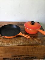 Photo showing both the orange enameled cast iron lidded pot and frying pan on wooden surface, handles and enamel loss visible.
