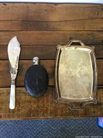 Front view of vintage knife with mother of pearl handle, leather covered flask, and Art Deco silver plate tray with engraved pattern on front.