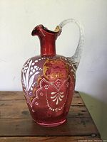 Front view of cranberry glass pitcher showing painted white floral decoration, gilded scrollwork, pontiled bottom, and clear twisted handle.