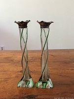 Pair of tall slender glass bud vases with green twisted lines and sterling silver ruffled rims against a white background on wood surface.