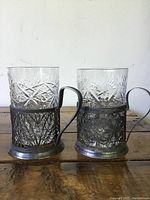 Front view of two Russian silver filigree cup holders with crystal cups inside on wooden surface.