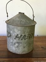 Front view of rustic, weathered metal oil tin with faded 'Ted Harrison' painted lettering, conical top, and wire handle.