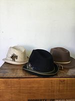 Three vintage men's hats placed side by side on a wooden surface with a white wall background. Hats are beige straw, black felt, and brown felt.