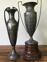 Both antique silver plate trophies shown together on wood surface, exhibiting overall wear and patina.