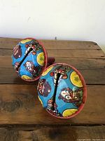 Pair of vintage clown rattles sitting on wooden surface, showing side angle and colorful stenciled clown faces with central handle