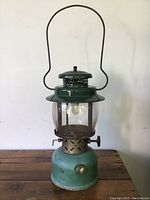 Full view of vintage Coleman green metal lantern showing the entire lantern including handle, glass globe, and metal base with visible surface wear and sun exposure.