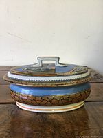 Side view of oval lidded soap dish showing blue and gold geometric painted details with rectangular handle on lid