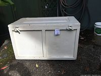 Photo showing the front side of the antique cream-colored wooden box on ground with metal latch locks.