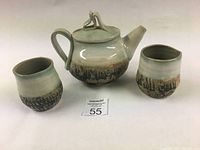 Photo showing the full teapot with handle, spout, lid, and two matching cups with textured design at the base and soft green-blue glaze at the top.