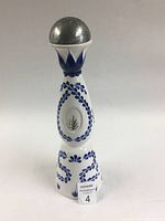 Front view of white ceramic tequila bottle featuring blue floral patterns and a metal cap, showing lot number tag
