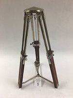 Front view of tri-pod stand with plumb bob showing metal legs with wood sections and central hanging plumb bob.