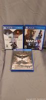 Three PS4 game cases including Killzone Shadow Fall, Mafia III, and Batman Arkham Knight arranged on a surface.