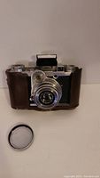 Front view of Zeiss Ikon camera mounted in brown leather half-case with removable metal lens cap placed beside.