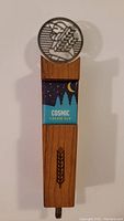 Front view of beer tap handle showing wooden body with 'COSMIC CREAM ALE' label and round metal emblem with hand holding barley design