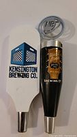 Beer taps photo showing both sides of the white Kensington Brewing Co. tap with blue and black logo and the black Nickelbrook Brewing Co. Headstock IPA tap with metallic NB logo on top.