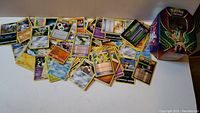 Wide view displaying a scattered assortment of assorted Pokemon cards alongside a Pokemon-themed collector's tin featuring Leafeon.