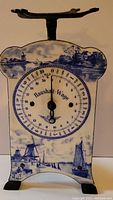 Full front view of the antique Dutch candy scale showing the entire porcelain body with blue hand-painted rural and windmill scenery and the dial with 'Haushalt-Wage' text.