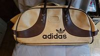 Front view showing Adidas logo in black with brown leather-like handles and trim against beige body of the bag