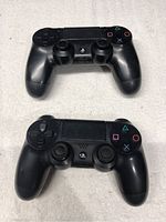Two black PlayStation DualShock 4 controllers placed on a white surface showing front view with buttons and sticks.
