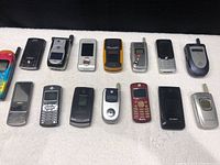 Lot of 15 older cell phones arranged in two rows on white cloth surface, showing the variety of flip and slide style phones in different colors and brands