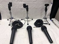 Full lot showing 3 black handheld microphones, 2 black audio cables, and 3 chrome tripod stands with black tape on clips.