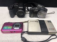 Four cameras placed on a white cloth: Fujifilm FinePix S1500, Panasonic Lumix DMC-ZS8, Fujifilm FinePix A pink compact, Konica Memrose 15 film camera