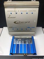 Pair of Lanzar audio amplifiers on a white surface, showing front tops and branding, one silver and one blue.