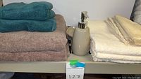 Shelf with stacks of teal, brown, and off-white bath towels with a ceramic soap dispenser, all neatly arranged.