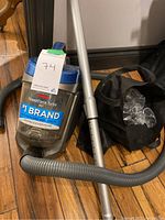 Bissell PowerForce Turbo Pet vacuum with metal extension tube, flexible hose, black accessory bag with plastic packaging inside on a wooden floor.