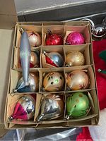 Opened box showing 12 vintage glass Christmas ornaments of various shapes and colors in cardboard dividers.