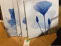 Four square blue and white floral canvas art pieces shown leaning against a wall on a wood floor