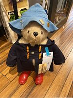 Front view of Paddington Bear showing blue felt hat with pins, navy blue duffle coat with wooden toggles, red plastic Wellington boots, and attached original cardboard tag.