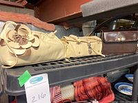 Lot of three handbags displayed on a black metal shelf, shows two beige bags on the left and center and a brown boxy handbag on the right.