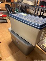 Two stacked plastic storage bins, beige with blue lid on top, gray on bottom, showing surface wear.